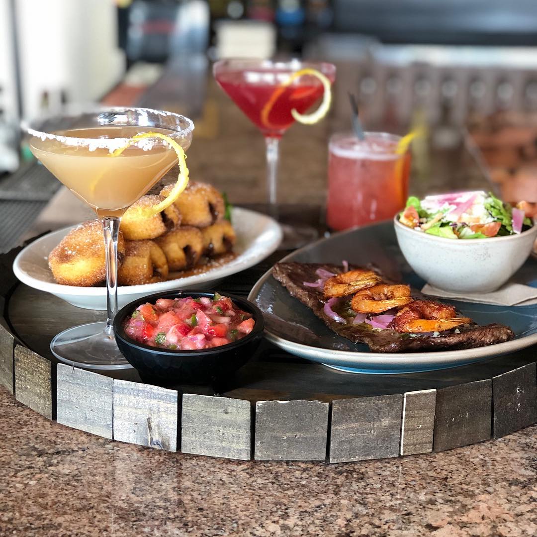 a plate with steak and a small bowl of salad. the second plate with small cinnamon donuts with three alcoholic beverages 